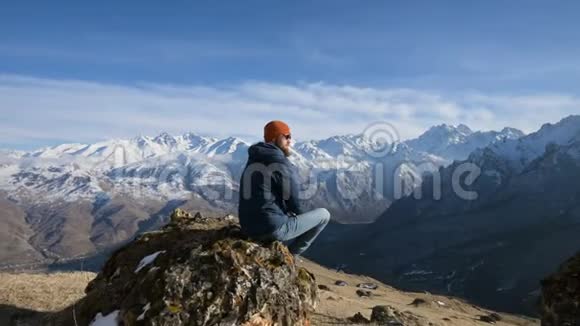 戴着太阳镜和帽子的留胡子旅行者的肖像坐在山峦背景下的岩石上视频的预览图