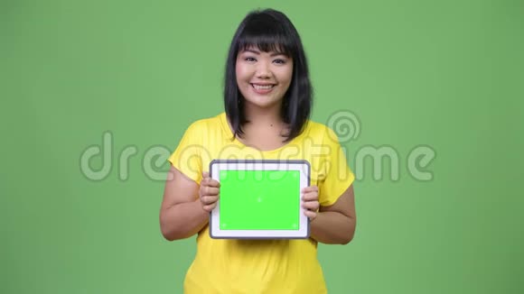 美丽快乐的亚洲女人一边思考一边展示数码平板电脑视频的预览图
