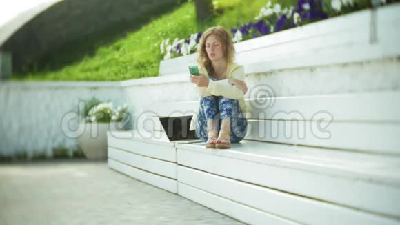 美丽的女人在公园的木凳上使用智能手机视频的预览图