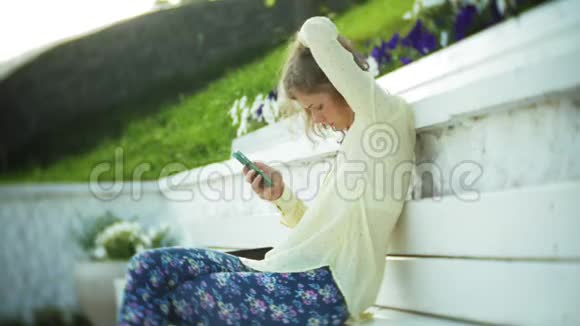 美丽的女人在公园的木凳上使用智能手机视频的预览图