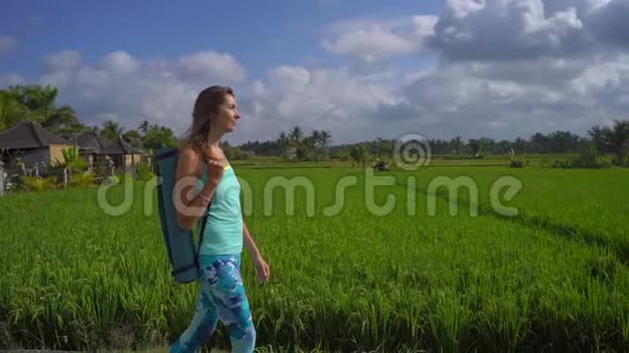 一个年轻女子用瑜伽数学在美丽的稻田里行走时的慢动作稳定镜头视频的预览图