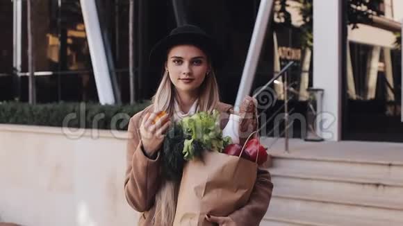 年轻漂亮的女人穿着时髦的外套站在街上手里拿着一包产品微笑着看着视频的预览图