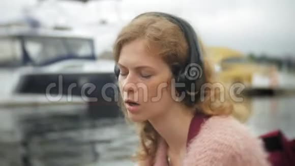 一个女孩在海滩上使用智能手机用耳机听音乐跳舞游艇在港口航行视频的预览图