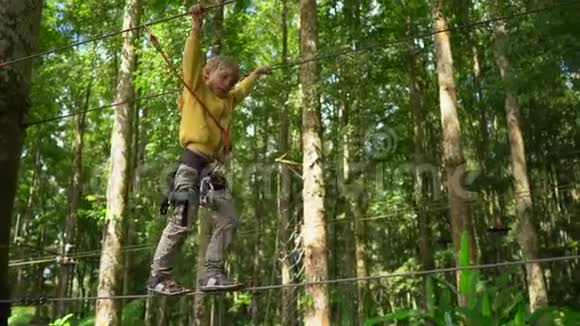 一个穿着安全装备的小男孩爬上森林探险公园的一条路线他爬在高绳上室外视频的预览图