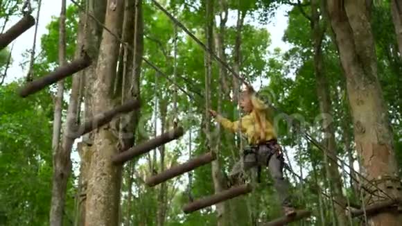 一个戴安全带的小男孩在森林探险公园的树梢上攀爬他在高绳道上攀爬视频的预览图