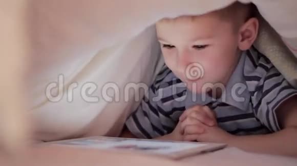 男孩躺在毯子下在触摸板上看卡通视频的预览图