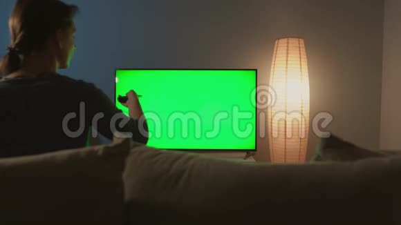 女人坐在沙发上用绿色屏幕看电视用遥控器切换频道彩色键视频的预览图