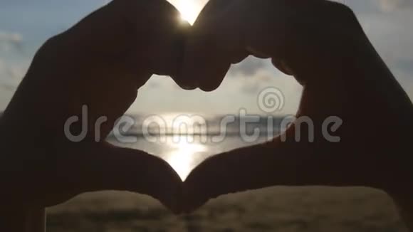 女孩用她的双手在海洋背景上用美丽的金色夕阳做心女性手臂的剪影视频的预览图
