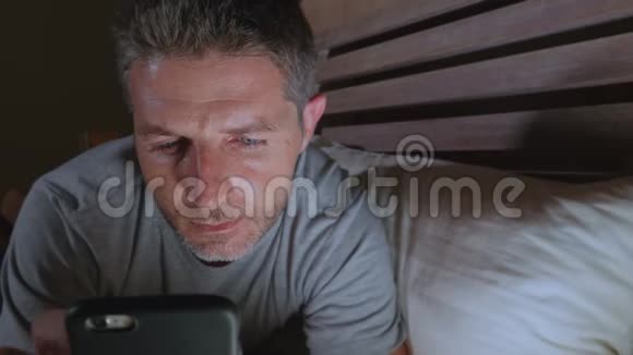 年轻迷人放松的男人深夜在黑暗的灯光下躺在床上用手机享受生活视频的预览图