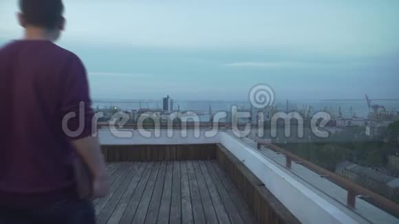 深色头发的男人穿着便服手里拿着日记看着阴天的海港海景走在木头屋顶上视频的预览图