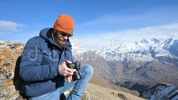 一位戴着太阳镜和帽子的长胡子旅行者摄影师的肖像坐在岩石上手里拿着镜子相机视频的预览图