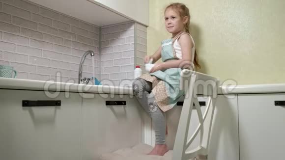 穿着围裙的漂亮小女孩站在椅子上在明亮的厨房里用海绵洗碗帮助视频的预览图
