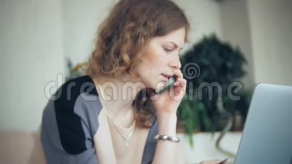年轻漂亮的商务女性坐在沙发上桌上放着平板电脑使用智能手机视频的预览图