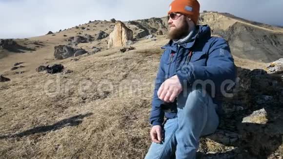 一位戴着太阳镜和帽子的留胡子旅行摄影师的肖像坐在岩石上手里拿着镜子相机视频的预览图