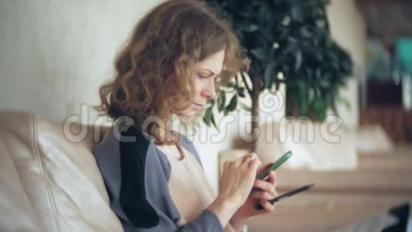 年轻漂亮的商务女性坐在沙发上桌上放着平板电脑使用智能手机视频的预览图