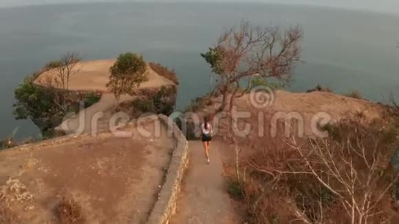 日出时女性在海边悬崖上慢跑的鸟瞰图视频的预览图