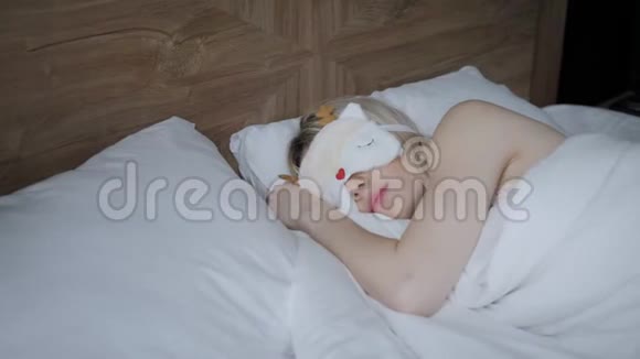 年轻女子睡在舒适的床上戴着面具睡觉眼睛上的眼罩早上在酒店房间白色枕头和视频的预览图