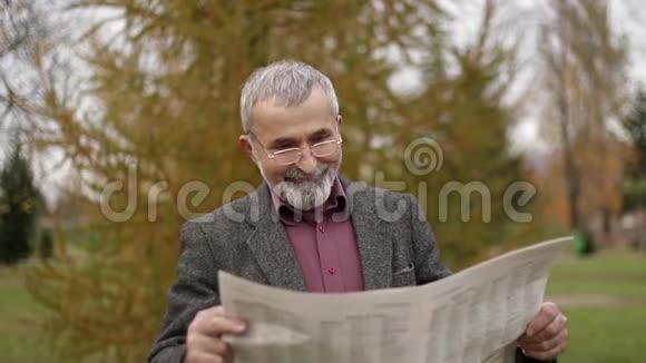 一位漂亮的祖父留着漂亮的胡子穿着灰色的夹克在公园里看报纸视频的预览图