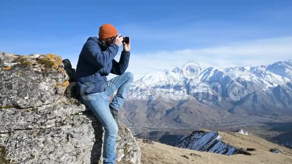 一位戴着太阳镜和帽子的长胡子旅行者摄影师的肖像坐在岩石上手里拿着镜子相机视频的预览图