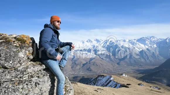 一位戴着太阳镜和帽子的留胡子旅行摄影师的肖像坐在岩石上手里拿着镜子相机视频的预览图