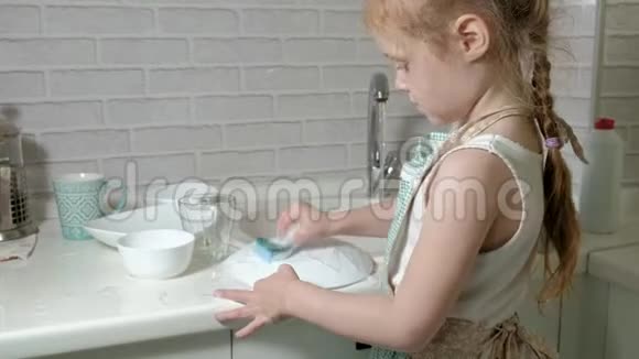 穿着围裙的漂亮小女孩站在椅子上在明亮的厨房里用海绵洗碗帮助视频的预览图