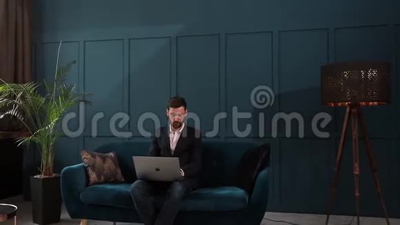 一位优雅的商人坐在豪华蓝色办公室内部的沙发上手提电脑的肖像视频的预览图