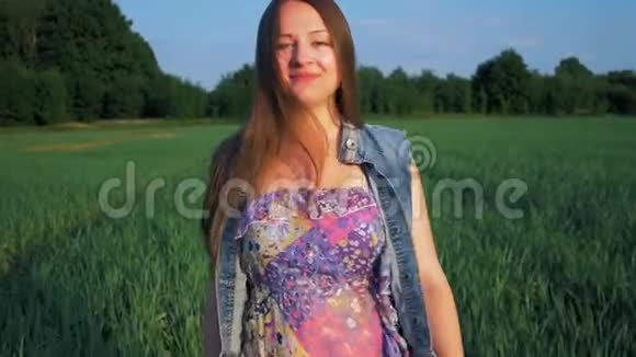年轻的女孩带着微笑直发沿着绿色的田野走着夕阳西下视频的预览图