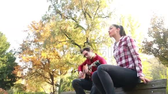 男人一边弹吉他一边女人唱歌坐在他旁边的公园长凳上视频的预览图