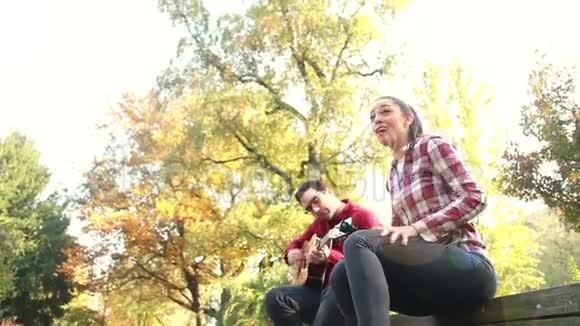 男人一边弹吉他一边女人唱歌坐在他旁边的公园长凳上视频的预览图