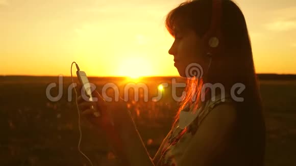 女孩在美丽的夕阳下听音乐跳舞戴着耳机和智能手机的年轻女孩视频的预览图