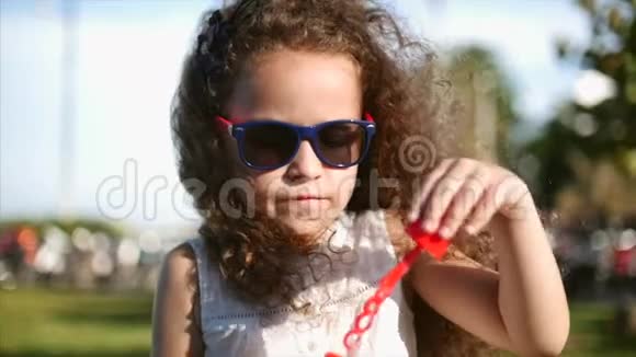 快乐的孩子在公园里吹肥皂泡漂亮的卷发小女孩戴着红眼镜吹着肥皂泡视频的预览图