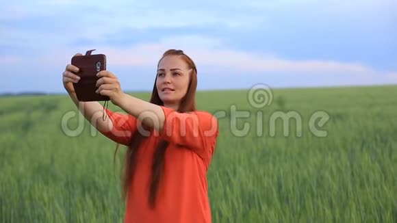 年轻的美女穿着红裙子红头发晚上在头发晚上在绿色的麦田打电话自拍视频的预览图