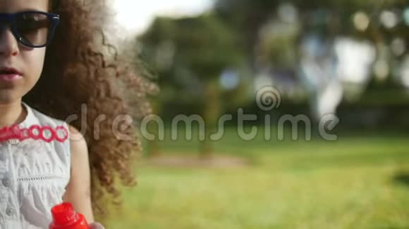 快乐的孩子在公园里吹肥皂泡漂亮的卷发小女孩戴着红眼镜吹着肥皂泡视频的预览图