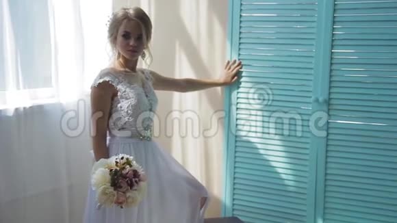 美丽的金发碧眼穿着一件白色的新娘礼服靠近一扇窗户放着一束鲜花视频的预览图
