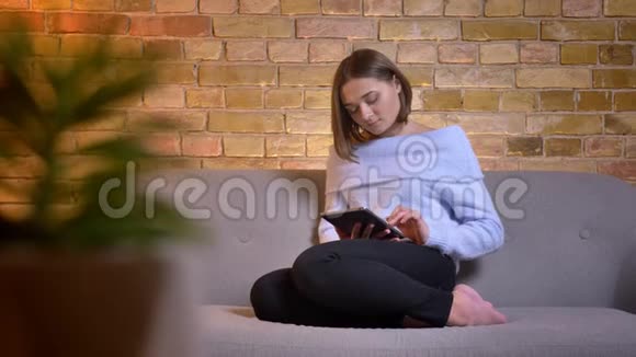 年轻美丽的白种人女性在舒适的家中坐在沙发上使用平板电脑的特写照片视频的预览图
