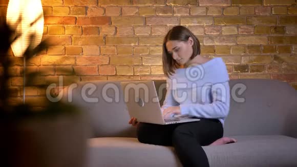 年轻漂亮的白种人女性在笔记本电脑上打字的特写镜头坐在舒适的室内沙发上视频的预览图