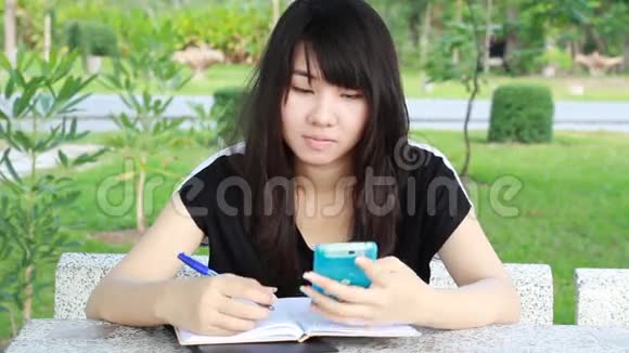 泰国学生十几岁的漂亮女孩用她的智能手机写了一本书坐在公园里视频的预览图