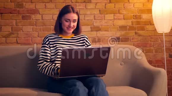 在舒适的室内公寓里年轻漂亮女孩用笔记本电脑坐在沙发上的特写肖像视频的预览图