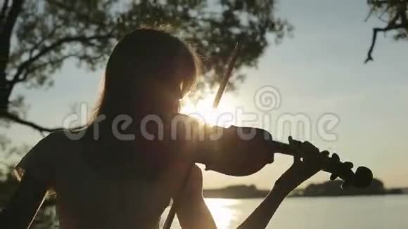 剪影女孩小提琴手在湖边日落时拉小提琴视频的预览图