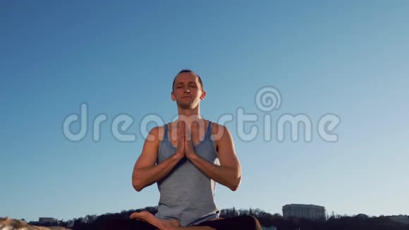 年轻的白种人通过在平静的河流附近的海滩上练习瑜伽健身运动放松身心背景是城市视频的预览图