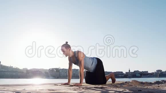 年轻的白种人通过在平静的河流附近的海滩上练习瑜伽健身运动放松身心背景是城市视频的预览图