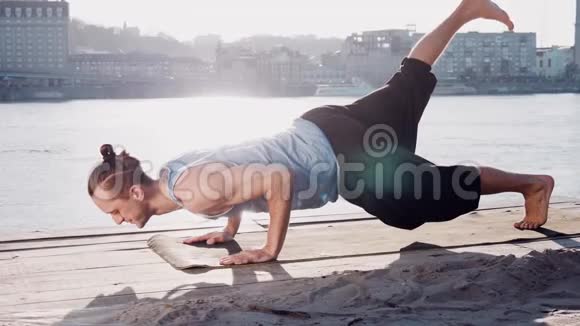 年轻的白种人通过在平静的河流附近的海滩上练习瑜伽健身运动放松身心背景是城市视频的预览图