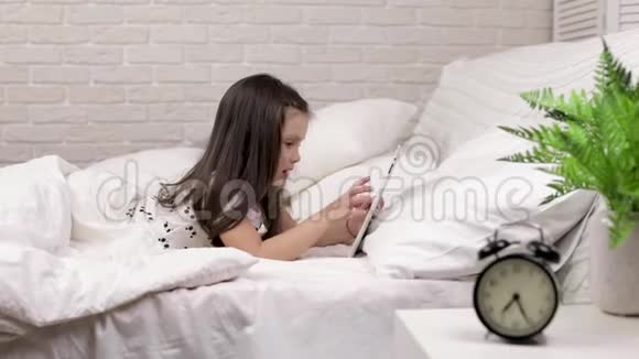 可爱的小女孩躺在床上使用数字平板电脑视频的预览图