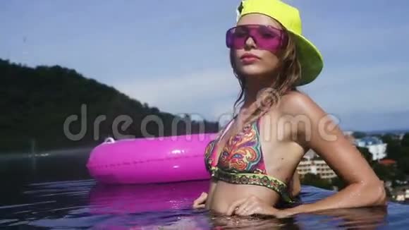 漂亮的年轻女人穿着比基尼黄色的帽子和紫色的太阳镜粉红色的充气戒指摆在无限的屋顶游泳池里视频的预览图