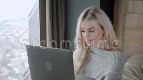 美丽的金发女人坐在床上带着灰色的铝制笔记本电脑女孩微笑心情好博客浏览互联网视频的预览图