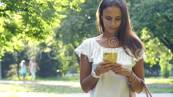 阳光明媚的一天一位迷人的年轻女子在夏天的公园散步时一边读写信息视频的预览图