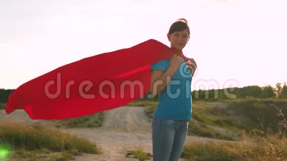 美丽的女孩超级英雄穿着红色斗篷站在田野上斗篷随风飘扬慢动作一个年轻的女孩视频的预览图