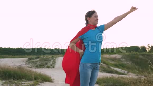 女孩梦想成为超级英雄美丽的女孩超级英雄穿着红色斗篷站在田野上斗篷飘扬在田野上视频的预览图