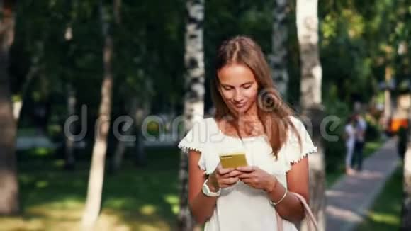 阳光明媚的一天一位迷人的年轻女子在夏天的公园散步时一边读写信息视频的预览图