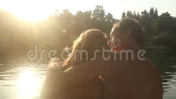 一对相爱的情侣在日落时坐在河边接吻和微笑视频的预览图